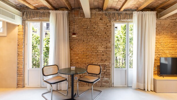 A cozy room with a brick wall, two windows with curtains, a round table with two chairs, and a TV on a stand. - Studio 2 pax in Born (Barcelona)