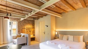 A modern hotel room with a large bed, a seating area with a sofa and armchair, a small table, and a stone wall. - Studio 2 pax in Born (Barcelona)