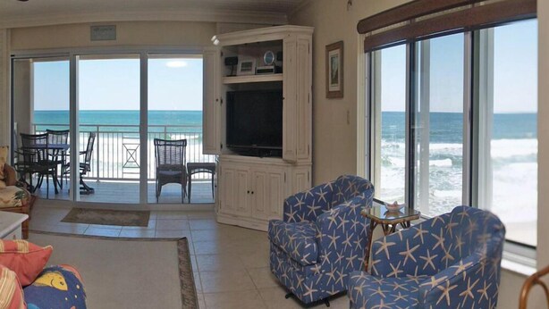 A spacious living room with a large TV, comfortable seating, and a view of the ocean through large windows. - Exclusive third floor three bedroom three bath end condo! (New Smyrna Beach)