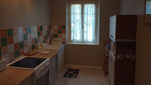 Une cuisine de taille réduite avec un mur recouvert de carreaux, une fenêtre ornée de rideaux en dentelle, une cuisinière et un réfrigérateur.