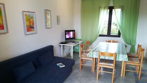 Ein Wohnzimmer mit blauem Sofa, einem Esstisch mit Stühlen, einem Fernseher und grünen Vorhängen.