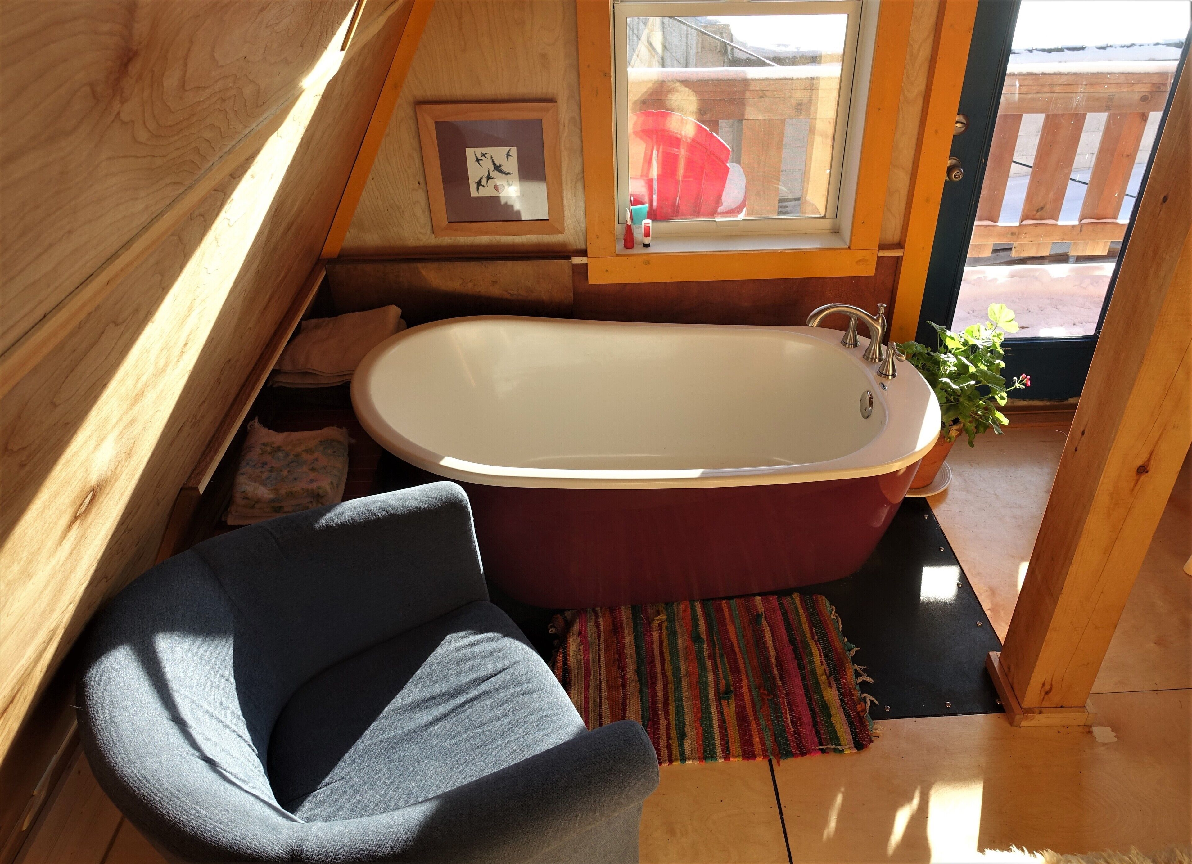 A small, cozy room with a wooden sloped ceiling, a single bathtub, a chair, and a window with a view of a wooden deck outside.