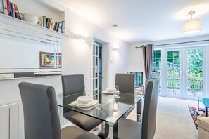 A modern dining area with a glass table, grey chairs, and a view of greenery outside.