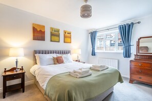 A hotel room with a bed, bedside tables, a dresser, a window with curtains, and artwork on the wall.
