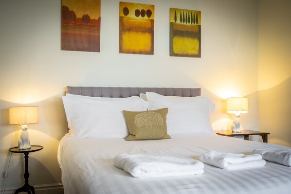 A neatly made bed with white linens and two pillows, a small round table with a lamp, and three abstract paintings on the wall.