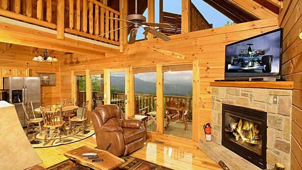 A wooden cabin interior with a fireplace, a television, and a balcony with a view of the mountains.