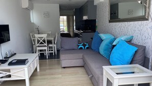 Un salon moderne avec un canapé gris, une table basse blanche et un téléviseur. On y trouve également un coin repas avec une table et des chaises blanches, et une cuisine à l’arrière-plan.