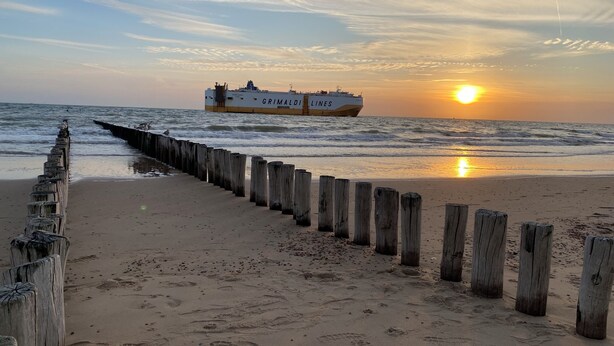 Une plage avec une jetée en bois et, au loin, un navire de la compagnie « Grimaldi Lines », le tout baigné par les couleurs du coucher de soleil.