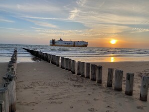 Una playa con un muelle de madera y un barco llamado 'Grimaldi Lines' al fondo durante el atardecer.
