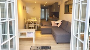 Un espace de vie compact comprenant un canapé, une table à manger et des chaises, un coin cuisine et une vue dégagée grâce à de grandes fenêtres.