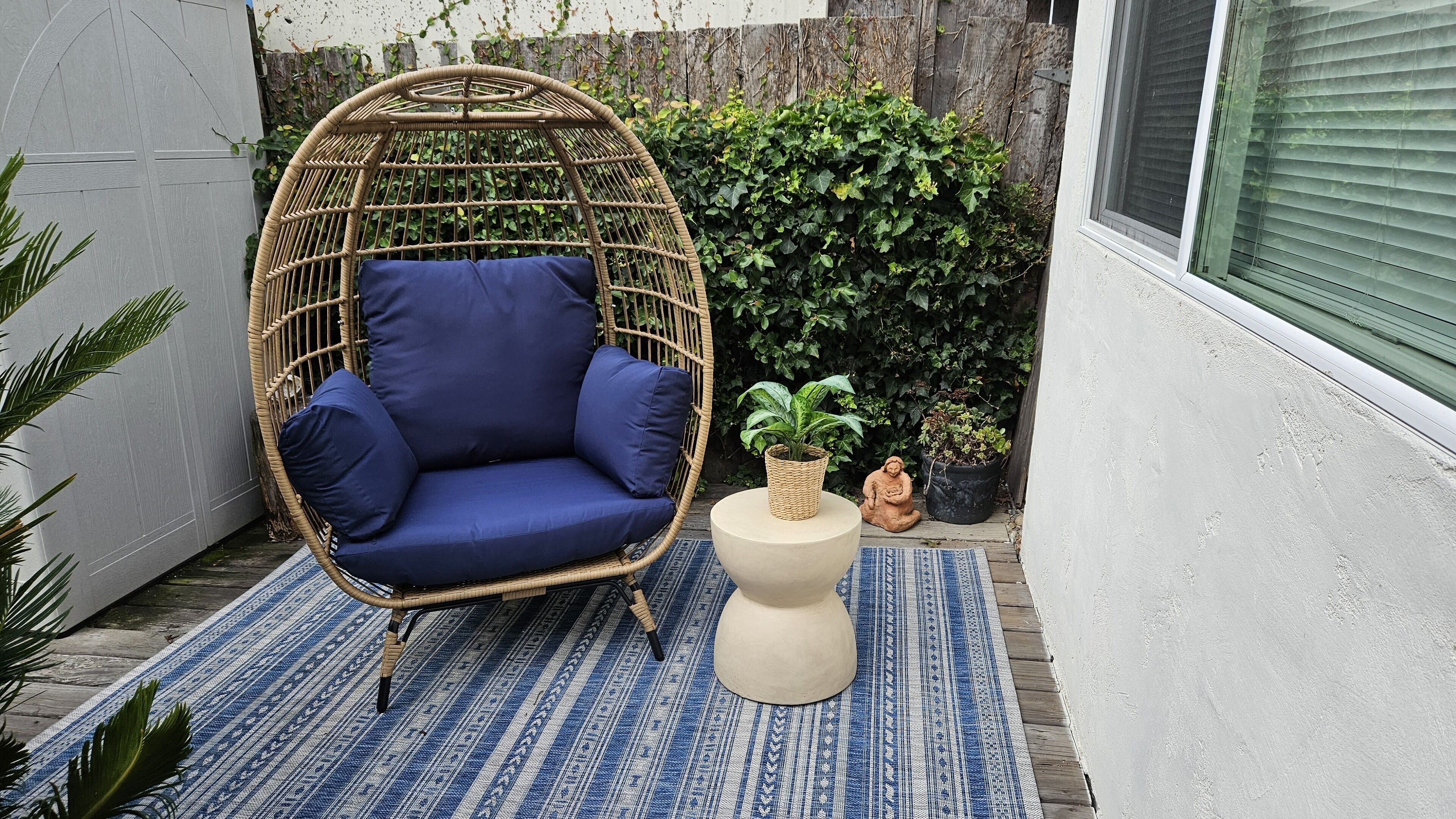 A wicker chair with blue cushions, a round table with a plant, and a patterned rug in a small outdoor space.