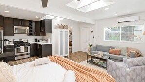 Un apartamento moderno con kitchenette, sala de estar con sofá y sillones, cama con manta, y un ventanal grande.
