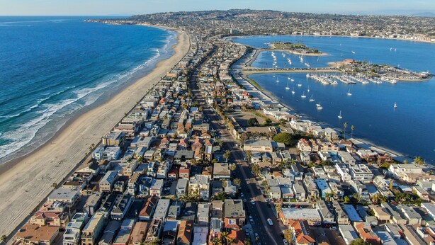 Vista aérea de un pueblo costero con una extensa playa, un puerto deportivo con barcos y edificios residenciales muy cercanos unos a otros.