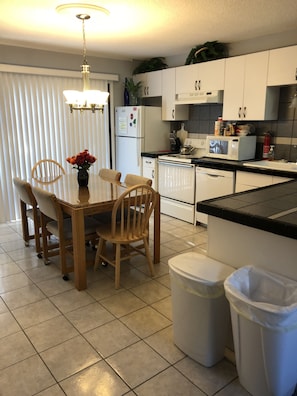 A kitchen with a dining table, chairs, a refrigerator, a microwave, and a sink. There are two trash bins and a ceiling light fixture.