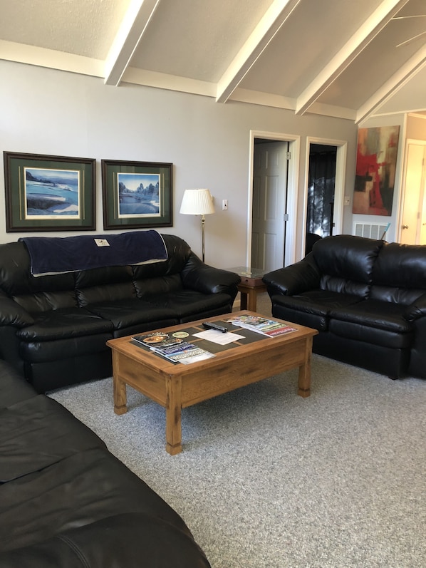A living room with two black leather sofas, a wooden coffee table with magazines, framed pictures on the wall, and a lamp.