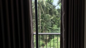 A room with a large window and a view of a balcony with greenery and a fence.