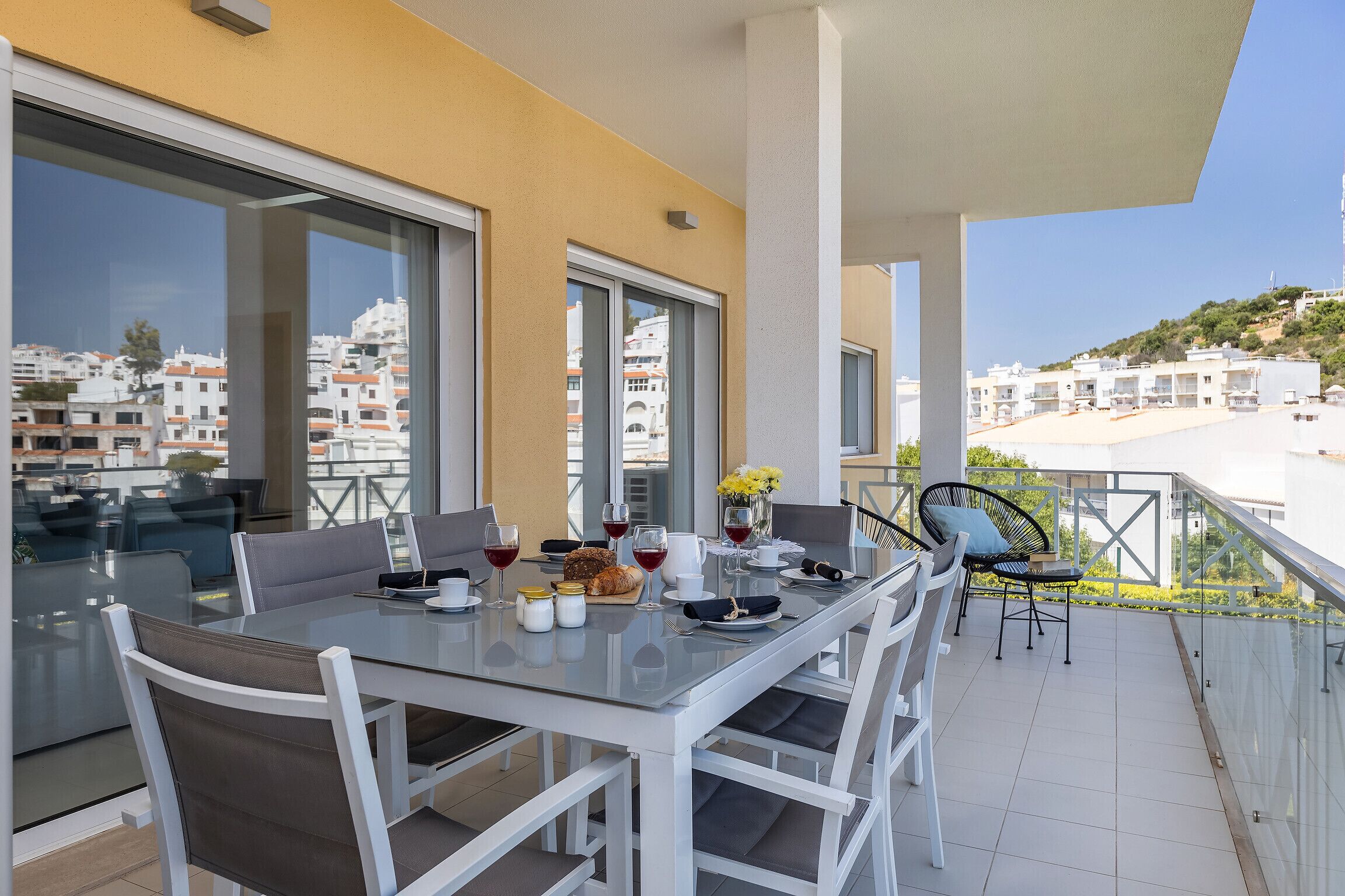Un coin repas avec une table en verre et des chaises blanches, un balcon offrant une vue sur les bâtiments et la plage, et une table dressée avec des verres à vin, du pain et des fleurs.