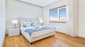 A modern bedroom with a large bed, a nightstand, a window with curtains, and a city view. - Flat Downtown OCV - 5min Beach (Albufeira)