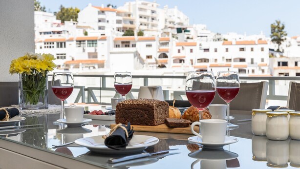 A table set with a loaf of bread, croissants, and a bottle of milk, with a view of white buildings in the background. - Flat Downtown OCV - 5min Beach (Albufeira)