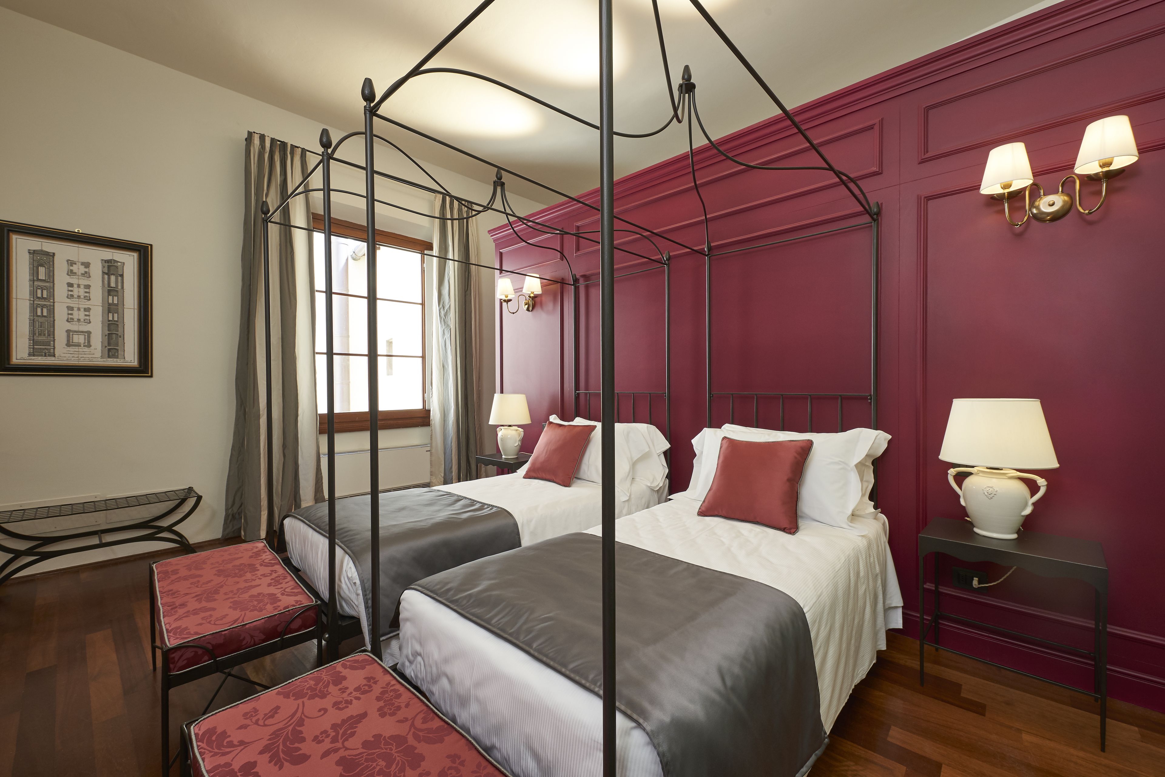 A four-poster bed with two single beds, a bedside table with a lamp, a framed picture on the wall, and a window with curtains.