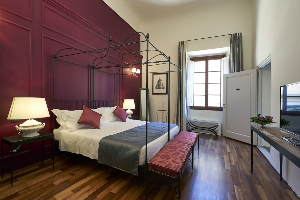 Ein Zimmer mit einem Himmelbett, einer Bank, einem Schreibtisch mit Computer und einem fenster mit Vorhängen.