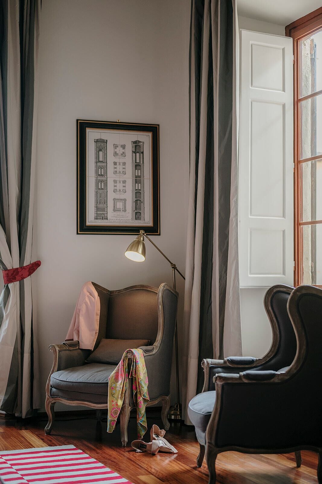 A room with a framed architectural drawing on the wall, a lamp, two chairs, and a window with curtains.