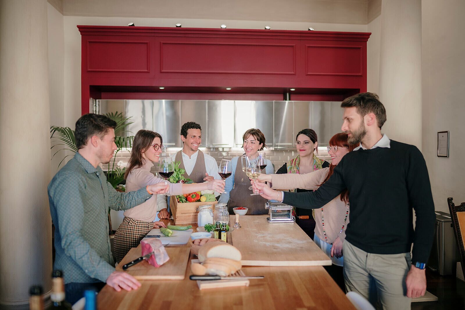 A group of people around a wooden table with food and drinks, in a room with red cabinets and a modern kitchen in the background.