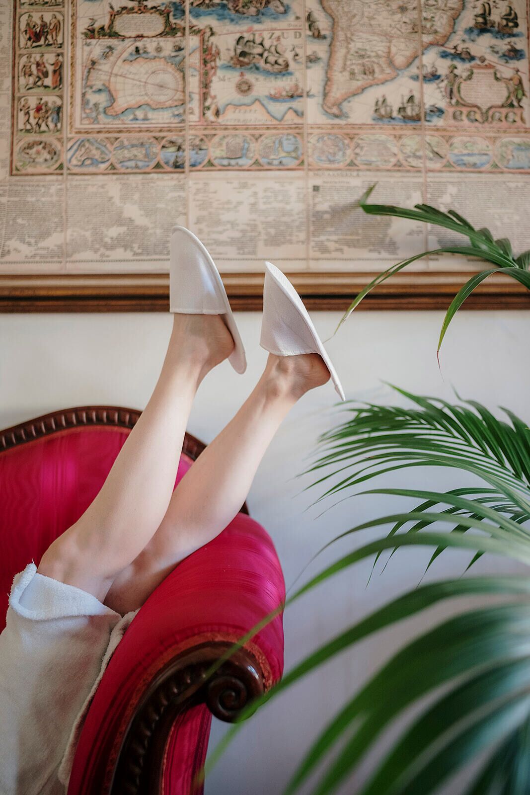 A person reclining on a red chair with white shoes, a large potted plant, and a wall with a detailed map.