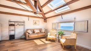 Une pièce spacieuse dotée d’un plancher en bois, d’un canapé beige, de deux fauteuils décoratifs, d’un porte-manteau en métal et d’une grande fenêtre offrant une vue dégagée.