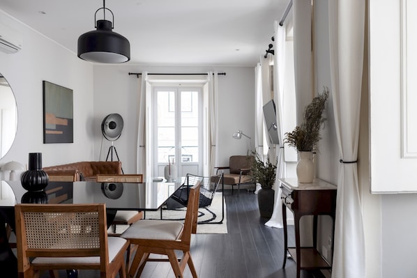 A modern living room with a dining area, featuring a sofa, chairs, a coffee table, and a large window with curtains.