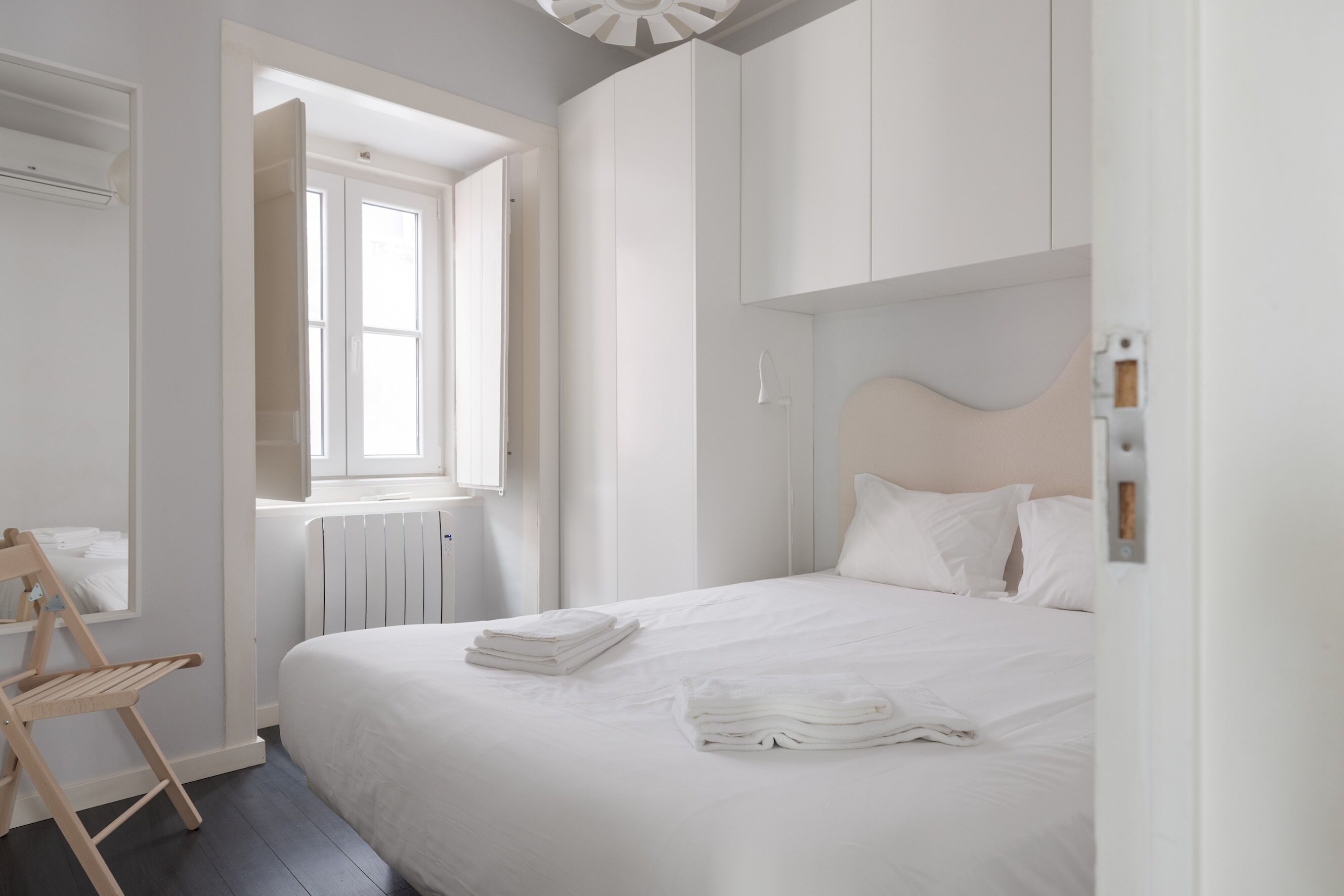Quarto com cama grande, roupa de cama branca, uma cadeira de madeira e janela com vista.