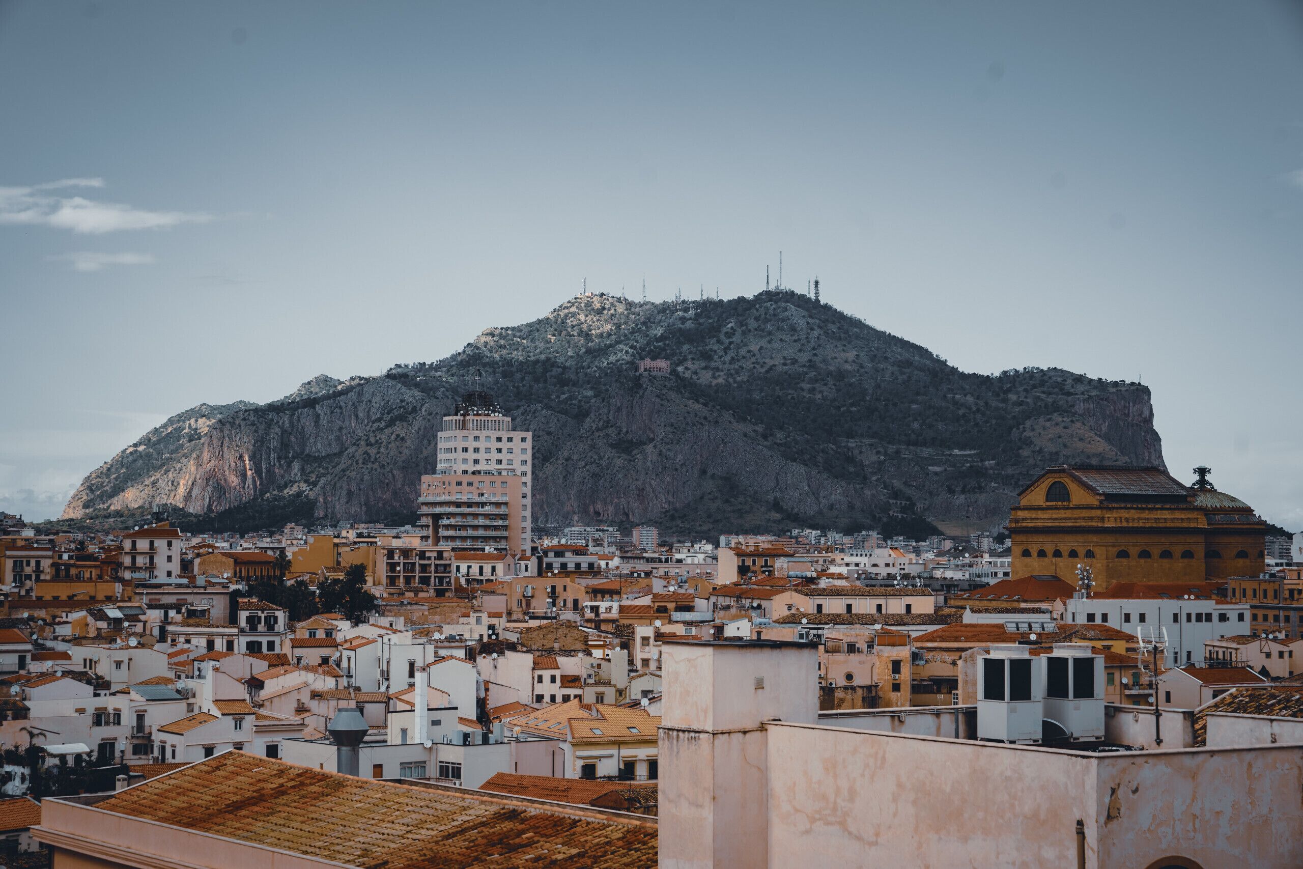 Un paysage urbain avec une montagne imposante en arrière-plan, un mélange de bâtiments résidentiels et commerciaux, et un bâtiment jaune remarquable surmonté d’un dôme.