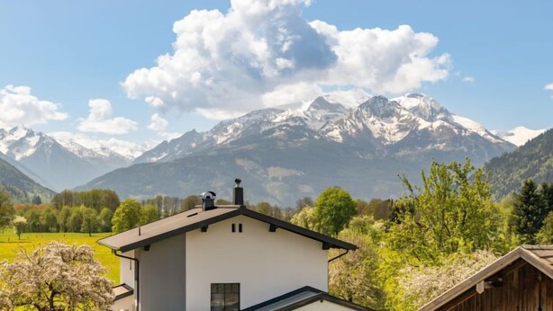 Une maison moderne avec un toit plat et une cheminée, entourée d’une végétation luxuriante et avec une chaîne de montagnes en arrière-plan.