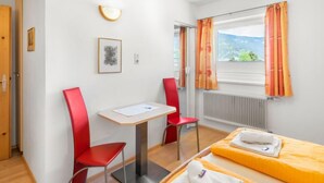 Une chambre d’hôtel avec un lit, deux chaises rouges, une petite table, un tableau encadré et une fenêtre avec des rideaux.