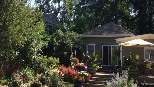 Ein Haus mit überdachter Terrasse, umgeben von einem Garten mit verschiedenen Pflanzen und Blumen.