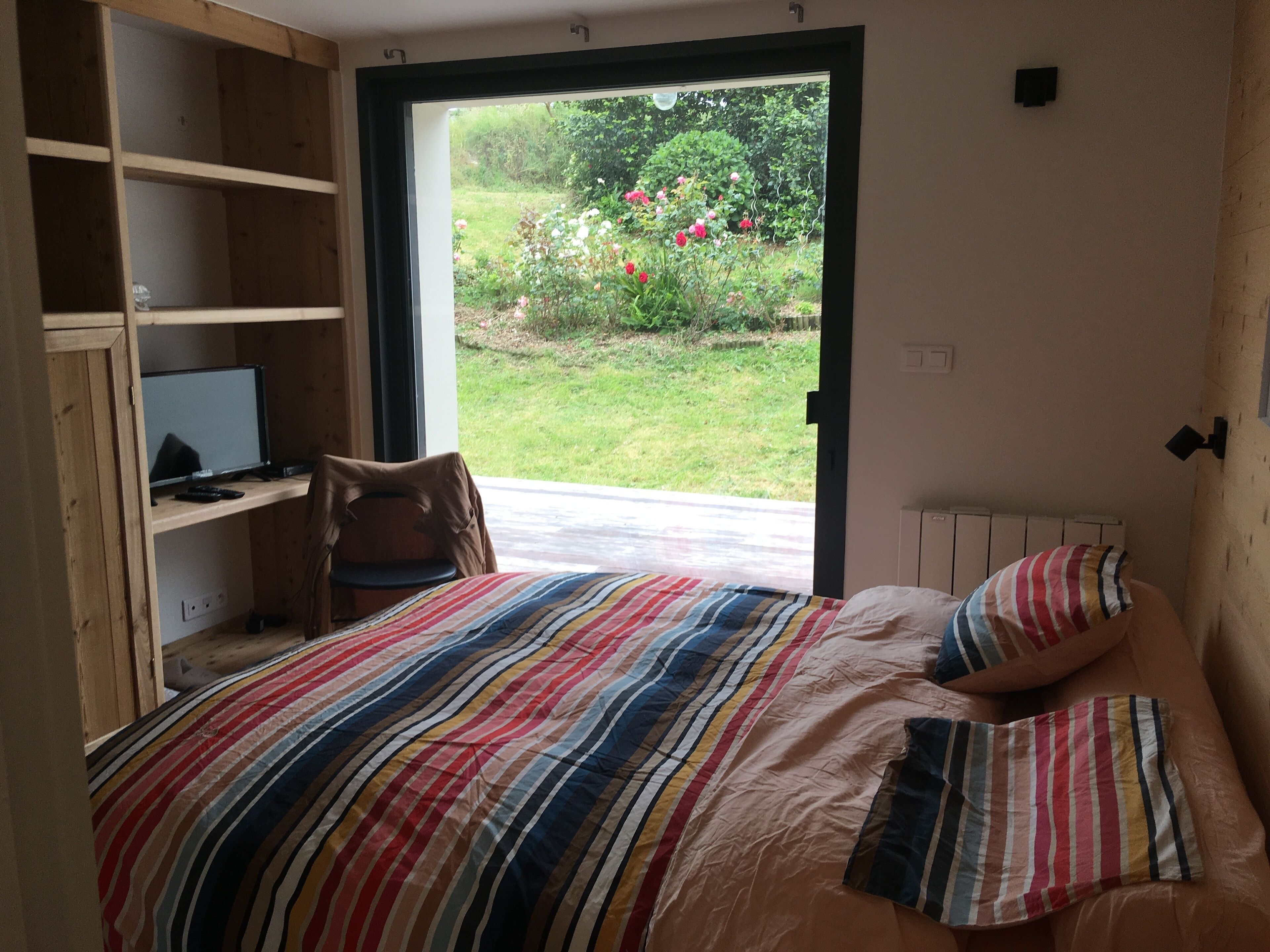 A bedroom with a bed, a chair, a desk with a computer, and a view of a garden.