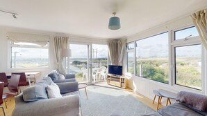 A spacious living room with a sofa, chairs, a TV, and large windows offering a view of the countryside. - E28 HI-Up, Riviere Towans (Hayle)
