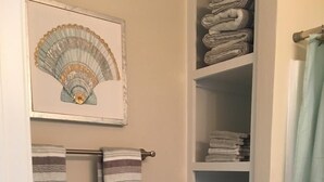 Une salle de bain avec des toilettes, un porte-serviettes avec des serviettes à rayures, une étagère avec des serviettes soigneusement pliées et une œuvre d’art encadrée au-dessus du porte-serviettes.
