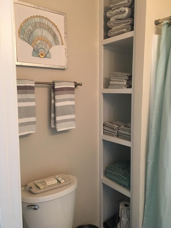 A bathroom with a toilet, a towel rack with striped towels, a shelf with neatly folded towels, and a framed artwork above the towel rack. - New Seaglass Cottage (Virginia Beach)