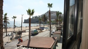 Vista costera con palmeras, una playa y un edificio de aspecto fortificado al fondo.