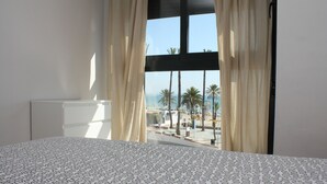 Habitación de hotel con cama, ventana con cortinas y vistas a la playa y palmeras.