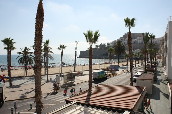 A coastal street with palm trees, a beach in the background, and a building with a red roof.