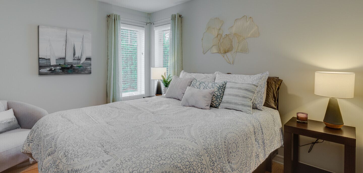 A bedroom with a large bed, a bedside table with a lamp, a chair, a window with curtains, and a wall art piece.