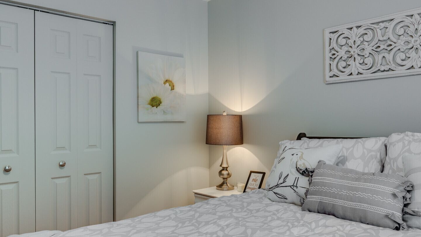 A bedroom with a bed, a nightstand with a lamp, a framed picture, and a decorative wall piece.