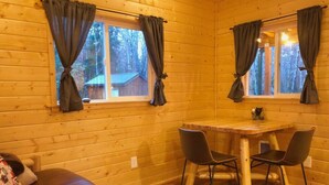 A cozy cabin room with a wooden interior, a bed, a dining table with chairs, and curtains on the windows.