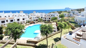A coastal resort with white buildings, a swimming pool, and a view of the sea and mountains.