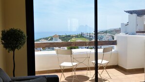 A balcony with a view of the sea and buildings. There are white chairs and a small table.