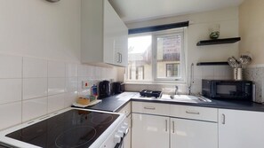 A compact kitchen with white cabinets, a black stove, a microwave, and a window with a view of buildings outside. - 15 Trevithick Court, Tolroy Manor (Hayle)