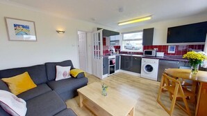 A compact living space with a sofa, a coffee table, a dining area with a table and chairs, a kitchen with a washing machine, and a TV mounted on the wall.