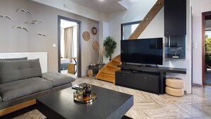Een moderne woonkamer met een grijze bank, een bijzettafeltje met een plant, een flatscreen televisie en een trap die naar een hoger gelegen verdieping leidt.